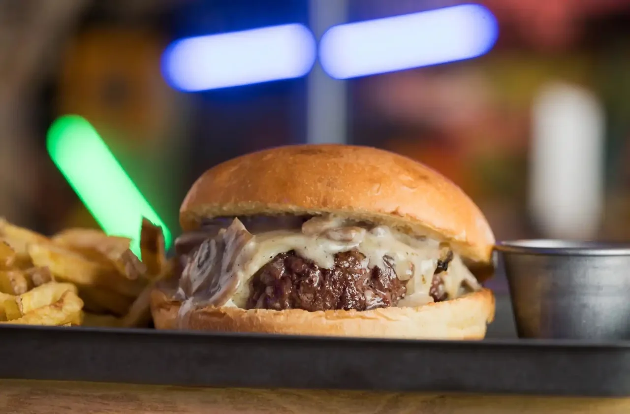 A burger with a melted cheese and mushroom topping on a tray beside fries and a small sauce pot