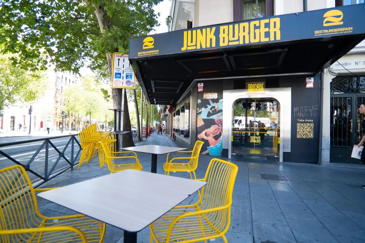Storefront exterior with a dark blue awning reading JUNK BURGER in yellow letters, yellow wire cafe chairs and tables on the sidewalk terrace.