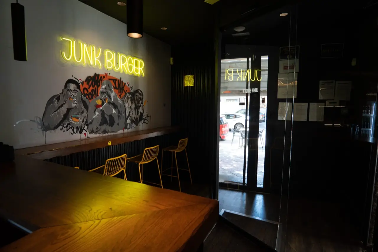 Interior bar counter with yellow stools, a painted gorilla mural, and a yellow neon sign reading JUNK BURGER on the wall, entrance door visible with reversed signage.