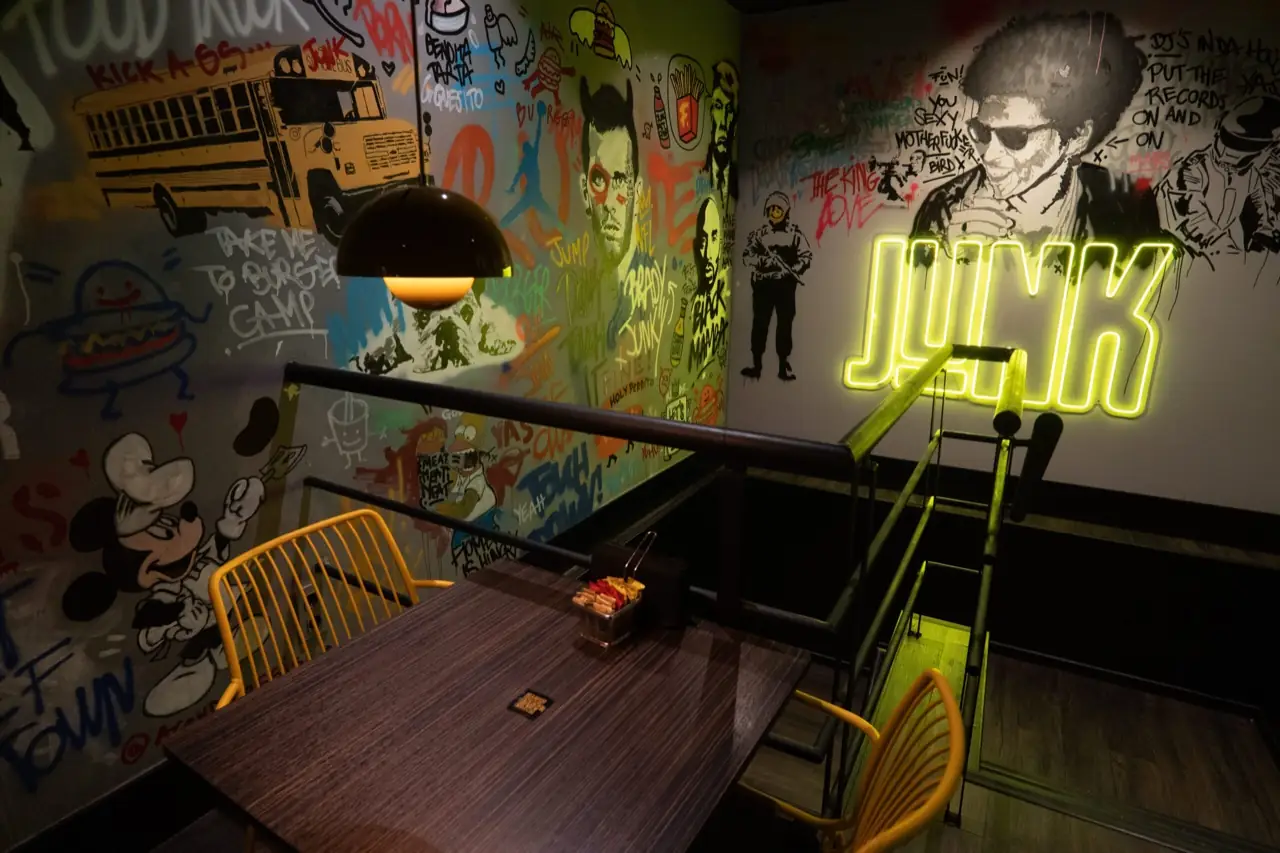 Restaurant interior seating nook with graffiti-style mural walls, a wooden table, yellow chairs and a yellow neon sign reading JUNK on the wall near a staircase.