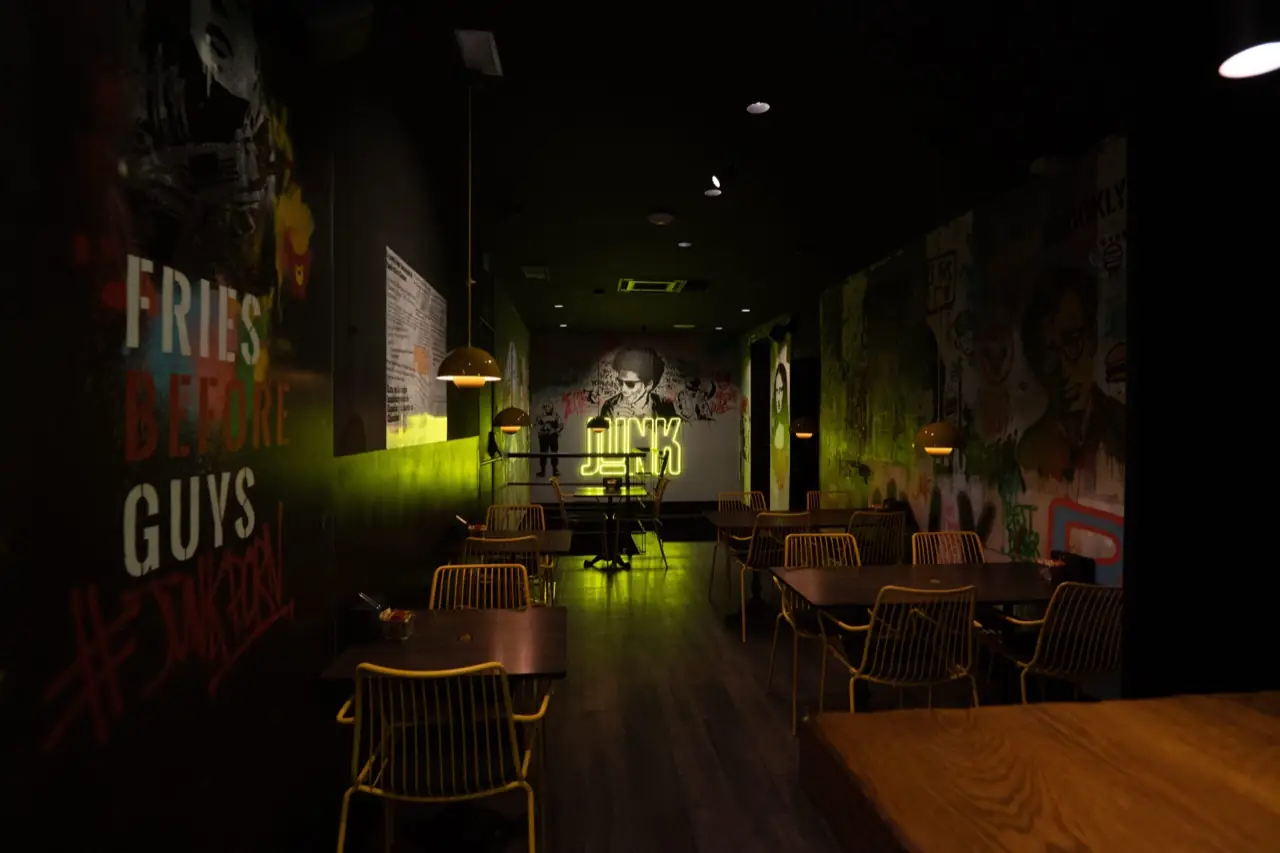 Dim restaurant interior dining room with graffiti-mural walls, wooden tables and yellow chairs, a yellow neon JUNK sign glowing at the far end.