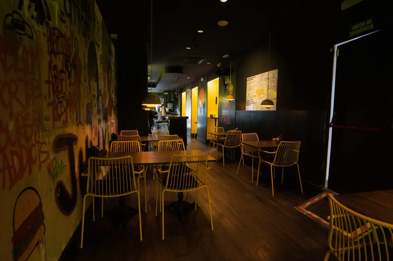 Restaurant interior dining room with graffiti-covered walls, wooden tables, yellow chairs, pendant lamps and an open emergency exit door.