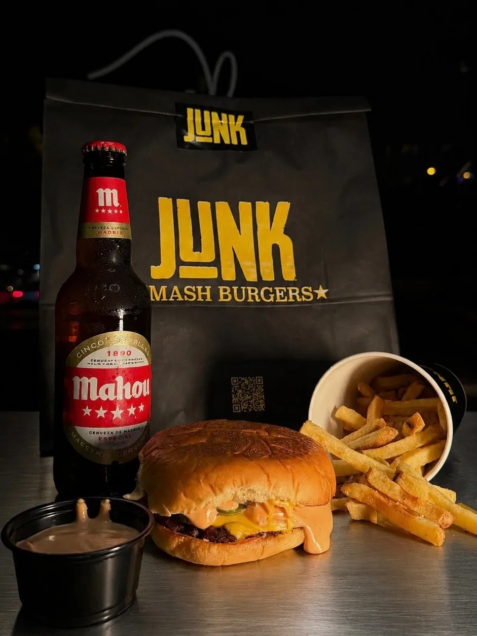 A cheeseburger with a cup of fries, a dipping sauce and a beer bottle in front of a black paper bag