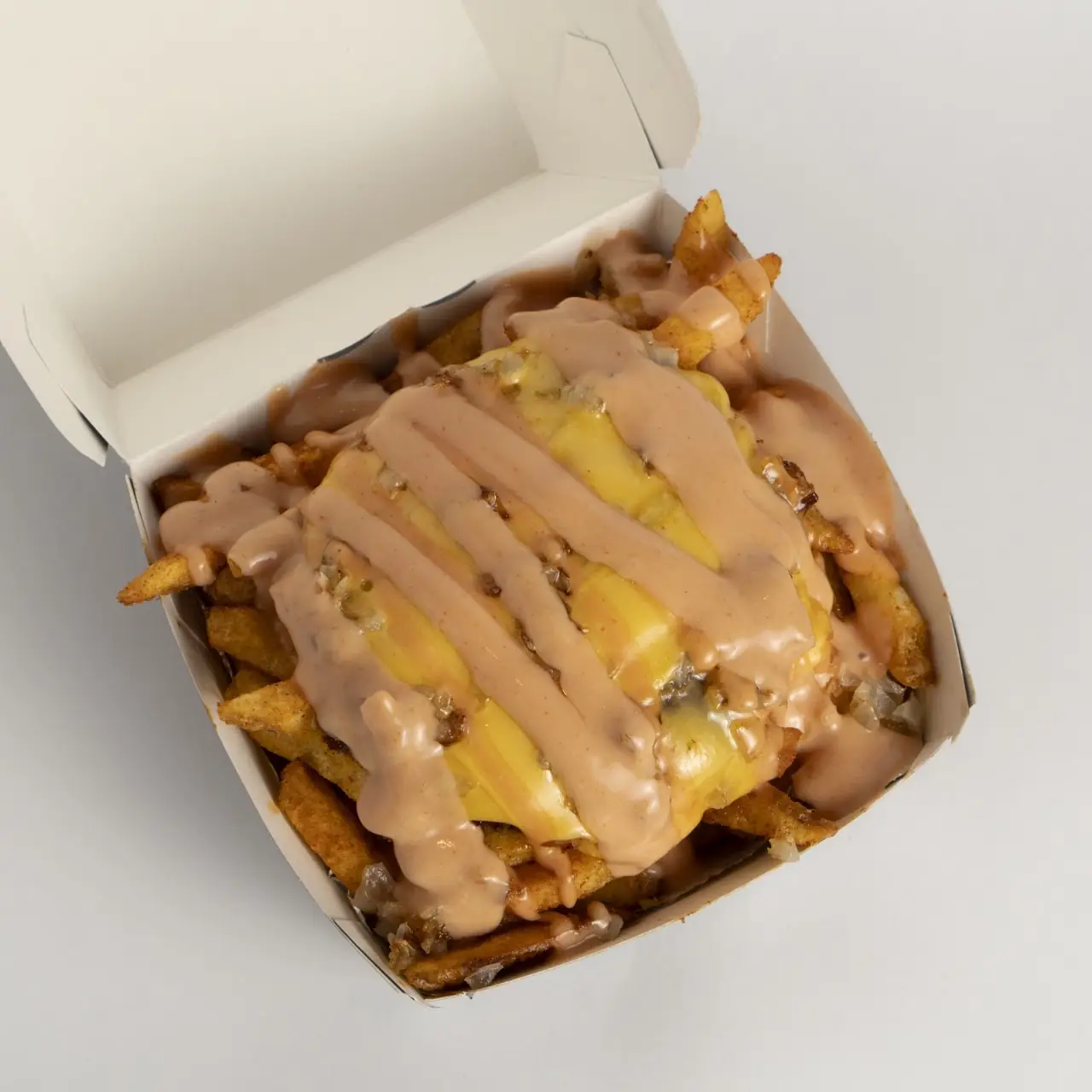 A takeout box of loaded french fries topped with melted cheese and a creamy sauce drizzle.