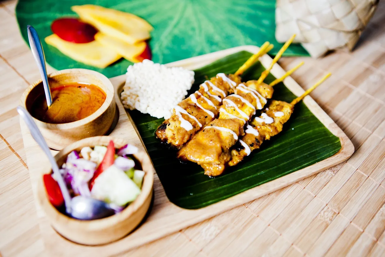 Grilled skewers with a drizzle of white sauce on a banana leaf beside bowls of dipping sauce and cucumber salad