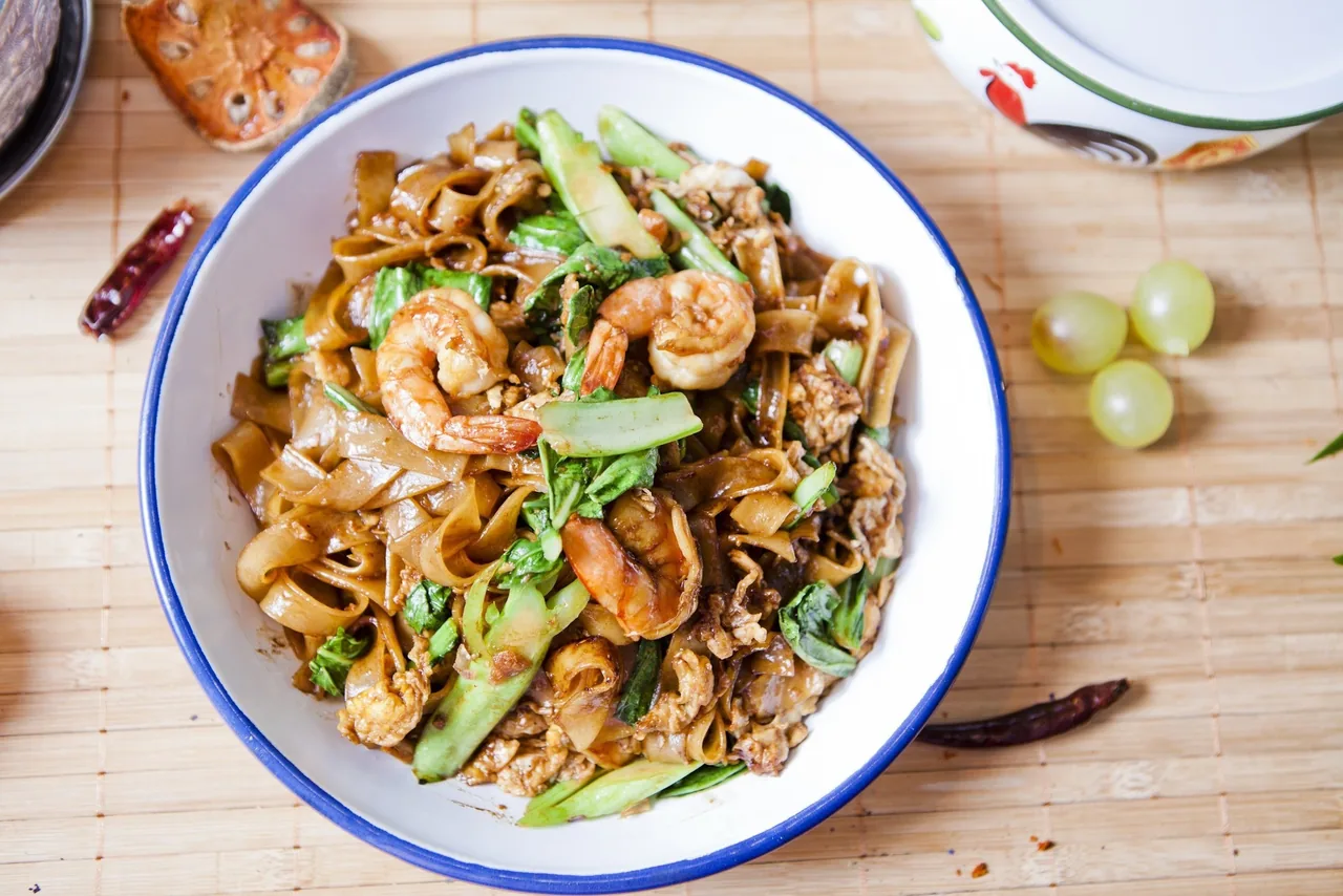 A white bowl of stir-fried wide noodles with prawns on a bamboo mat