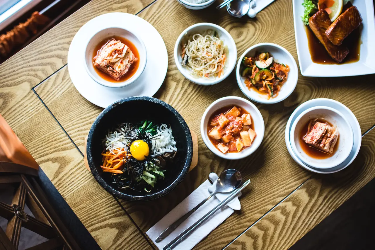 Overhead spread of Korean banchan bowls — kimchi, japchae, spicy cucumber, radish, and a stone-pot bibimbap topped with a raw egg yolk, carrots, and greens — at Kamasot