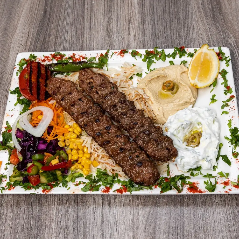 Two grilled kofte skewers over rice with hummus, tzatziki, lemon, and a fresh vegetable salad on a white plate, at Kapadokya