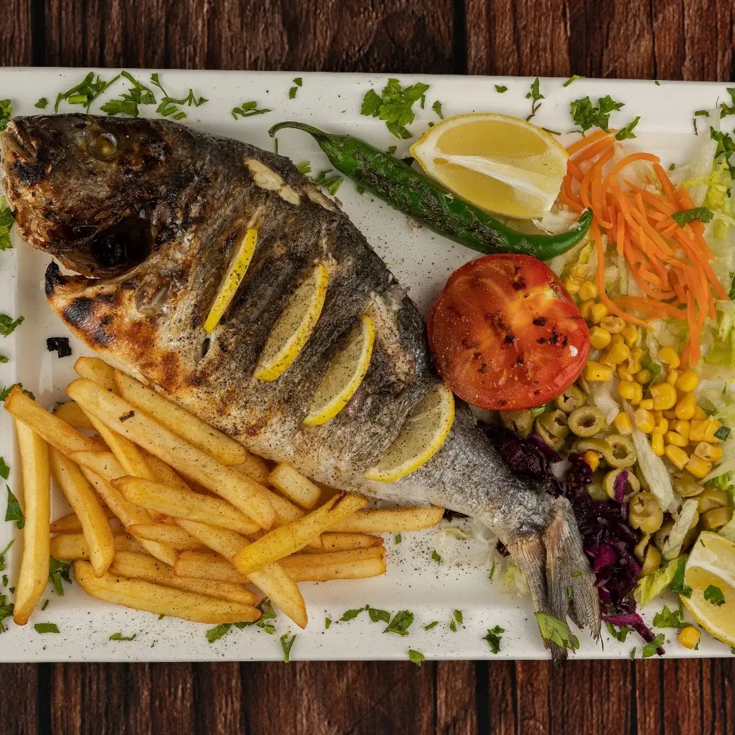 Whole grilled sea bream with lemon slices, fries, grilled tomato, and a fresh vegetable salad on a white plate, at Kapadokya