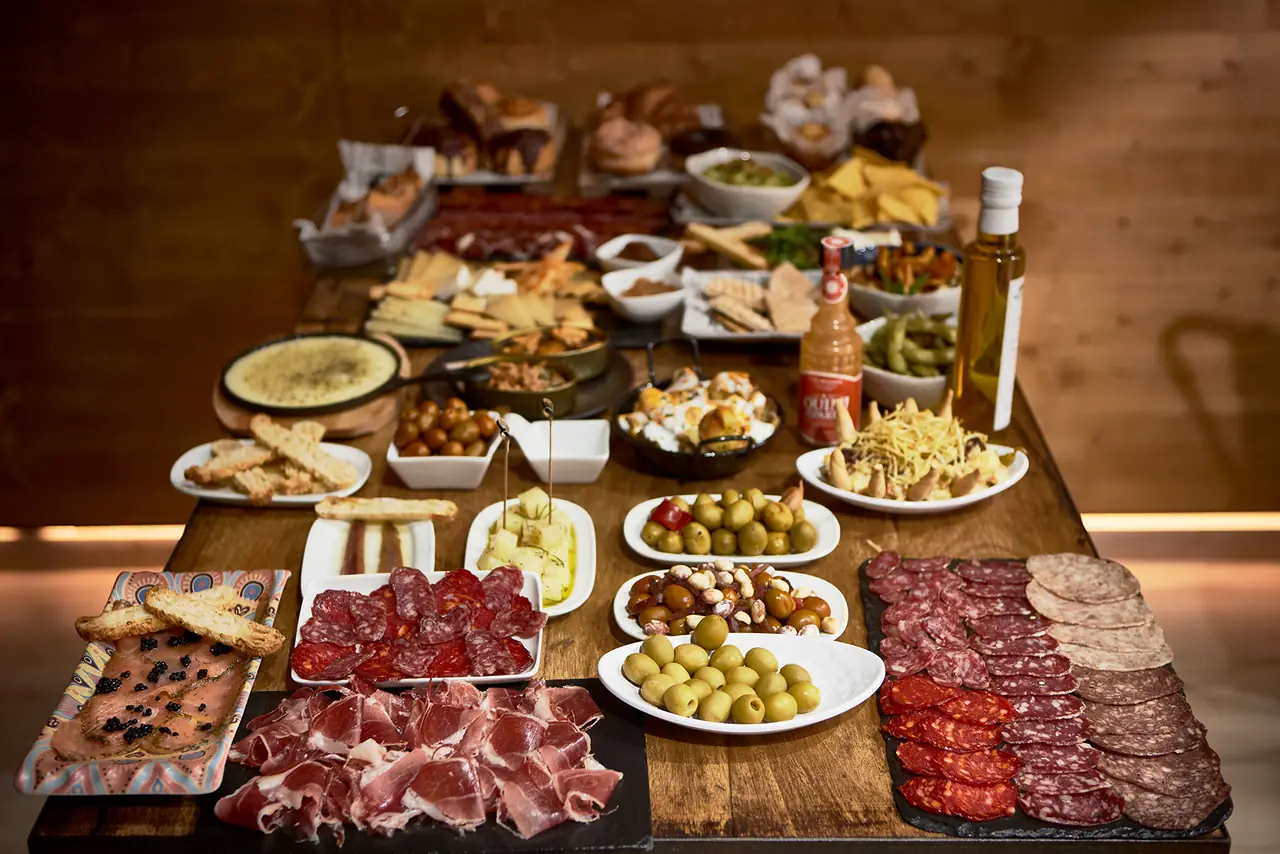 Spread of Spanish tapas on a wooden table, including sliced jamón and cured sausages, marinated olives, a cheese board, patatas bravas, croquettes, edamame, and toasted bread, at Karli