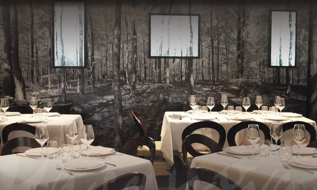 Dining room with white-clothed tables set with wine glasses, dark bentwood chairs, and a black-and-white forest mural covering the back wall with three lit vertical panels