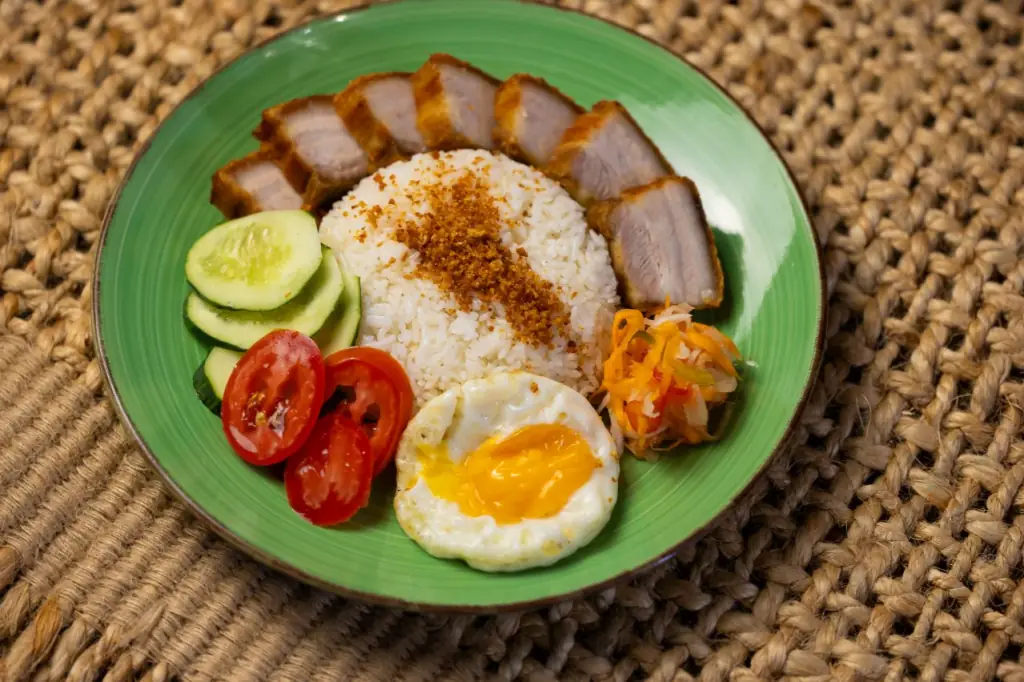 Rice plate on a green ceramic dish with sliced roast pork belly, a fried egg with runny yolk, sliced tomato, cucumber, and shredded pickled carrot, Kasarap Filipino Kitchen