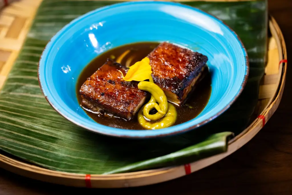 Two pieces of glazed, seared pork belly in a dark sauce with a swirl of yellow mustard, served in a blue bowl on a banana leaf, Kasarap Filipino Kitchen