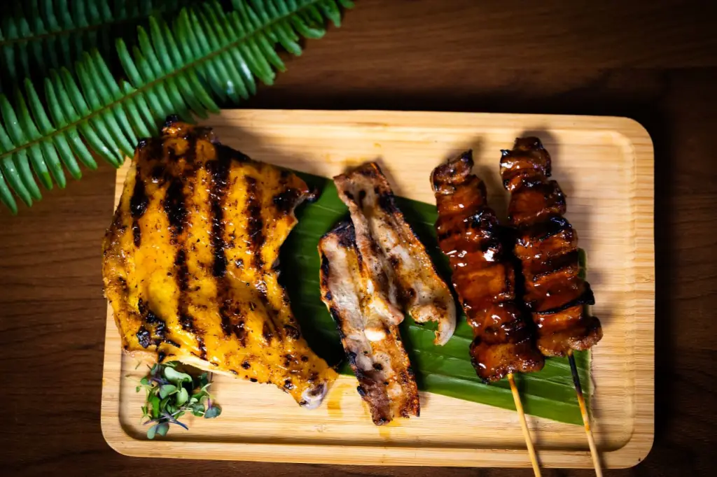 Grilled skewers and charred meat cuts arranged on a wooden board with a banana leaf, seen from above, Kasarap Filipino Kitchen