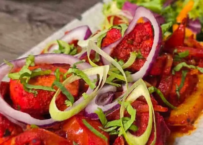 Tandoori chicken tikka pieces charred red, topped with red onion rings, leek curls and chopped cilantro on a platter