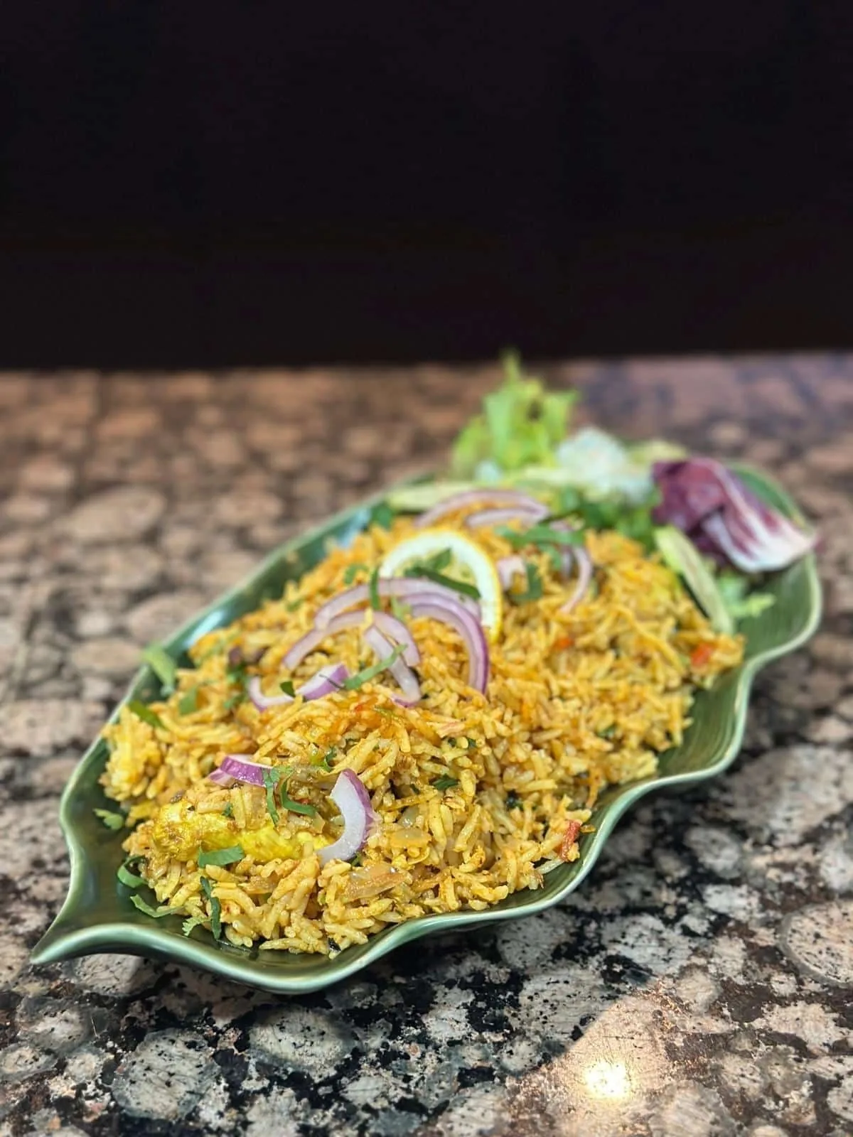 A green leaf-shaped plate of spiced rice with sliced red onion, lemon and salad leaves