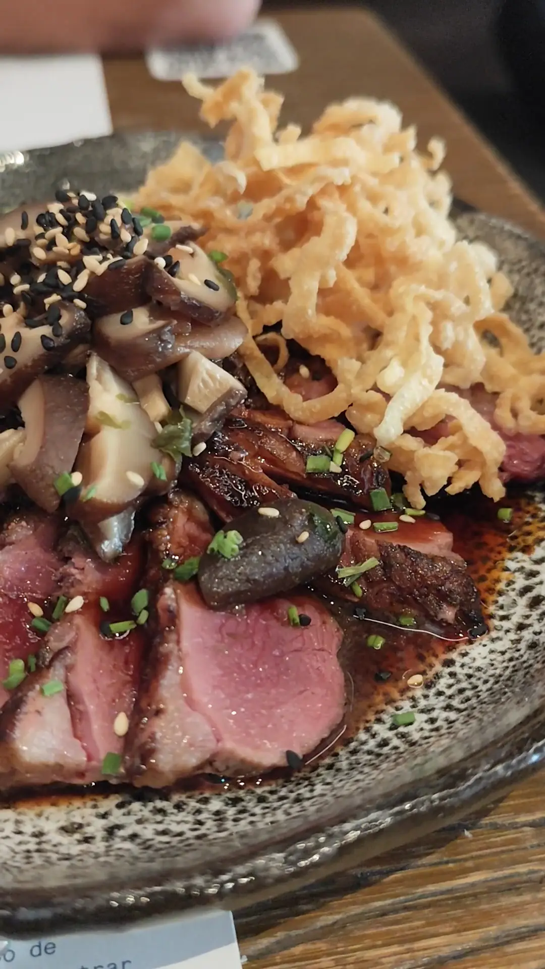 Sliced seared beef with crispy fried onions, mushrooms, and sesame seeds on a speckled plate.