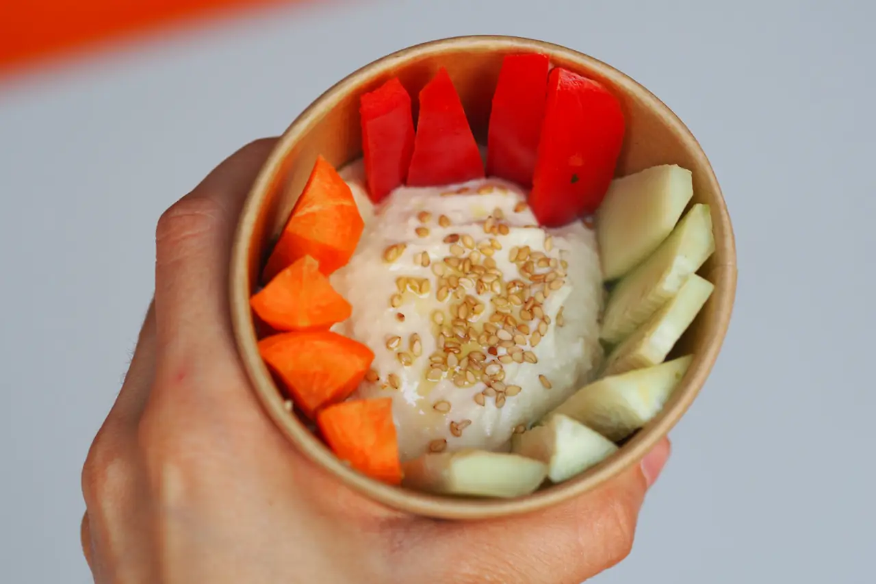 Takeaway bowl of hummus topped with sesame seeds and olive oil, served with sliced carrot, red pepper, and cucumber, Kata