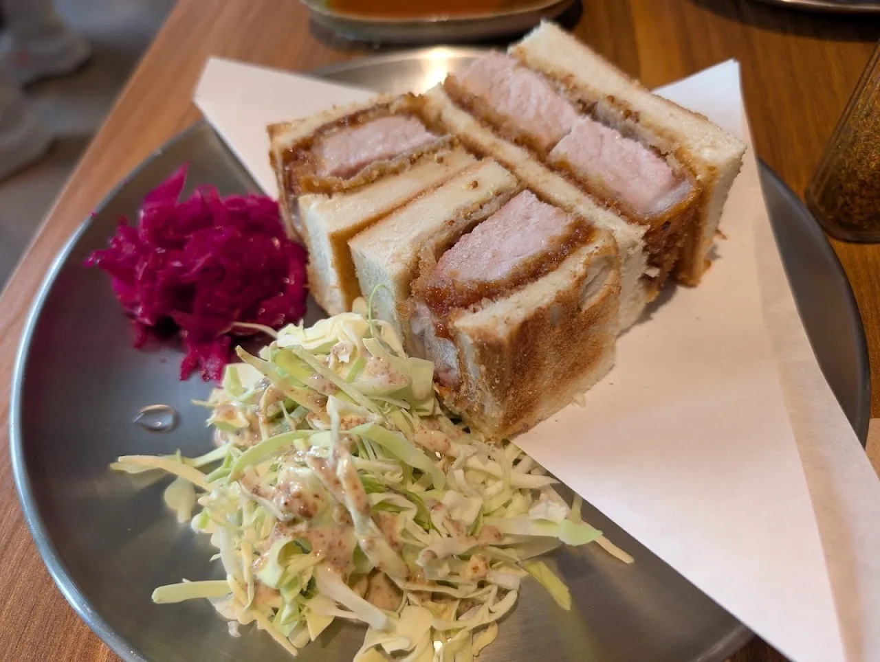 Cut sandwich halves filled with fried cutlet on paper, beside shredded cabbage and pickled red cabbage