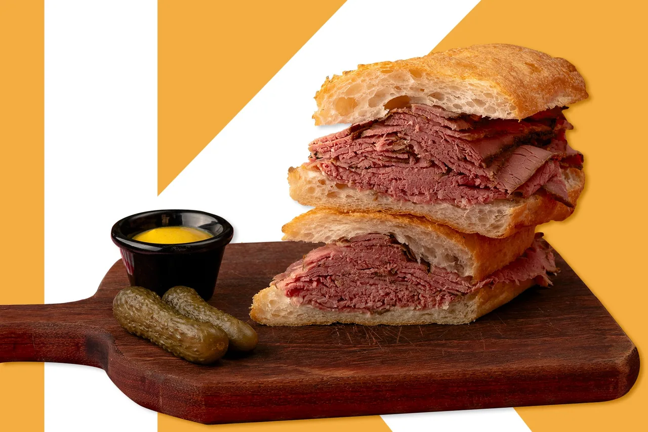 A pastrami sandwich on ciabatta cut in two and stacked on a wooden board, with two gherkins and a pot of mustard