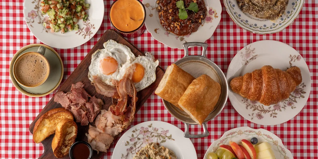 Overhead spread on a red gingham cloth: fried eggs, bacon and sliced pastrami on a board with fried bread, a chopped salad, a sweet onion dish, pastries, a croissant, sliced fruit, coffee and a glass of juice