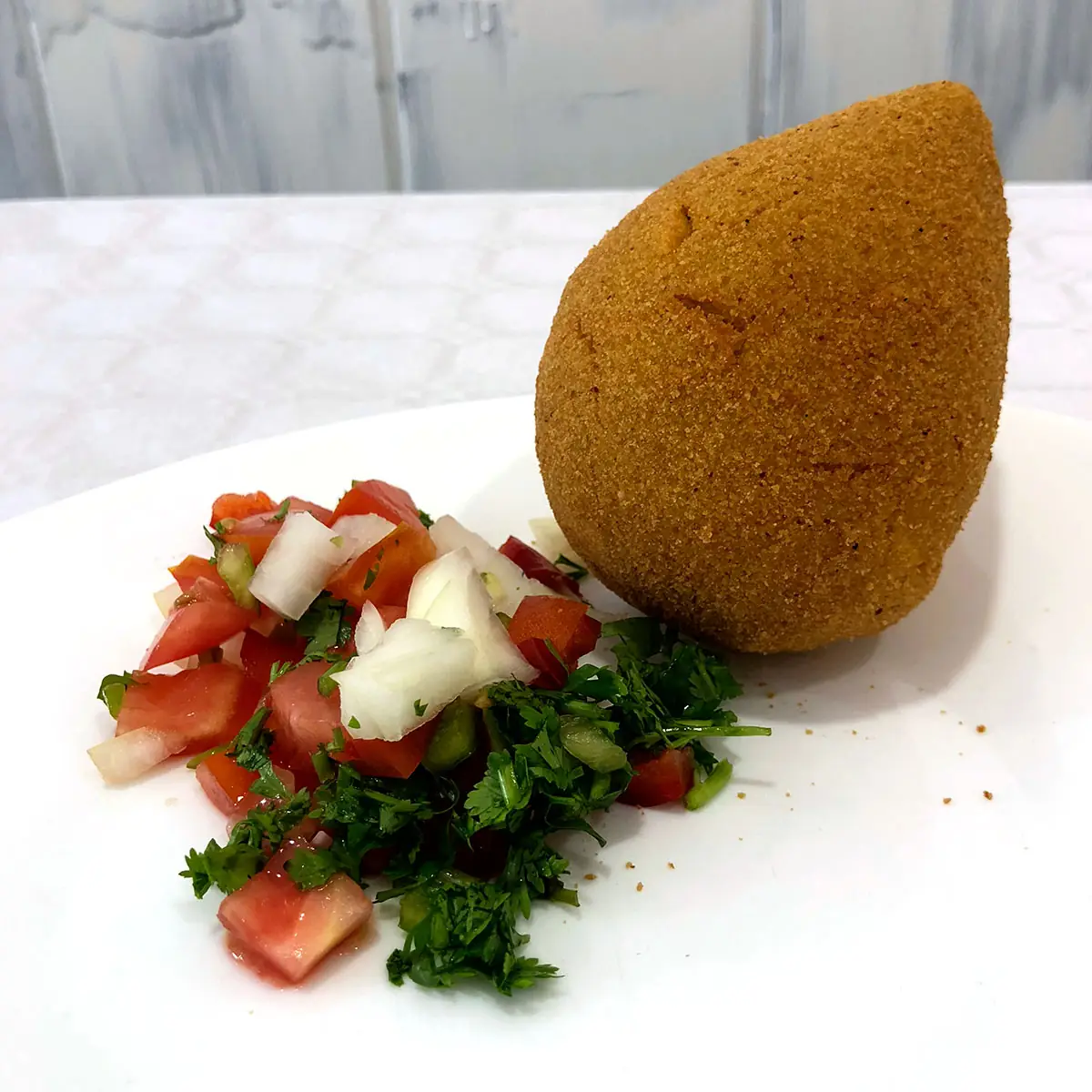Golden fried coxinha (Brazilian breaded chicken croquette) on a white plate with a side of chopped tomato, onion, and cilantro salsa, at K'Delicia Restaurant Brasileño