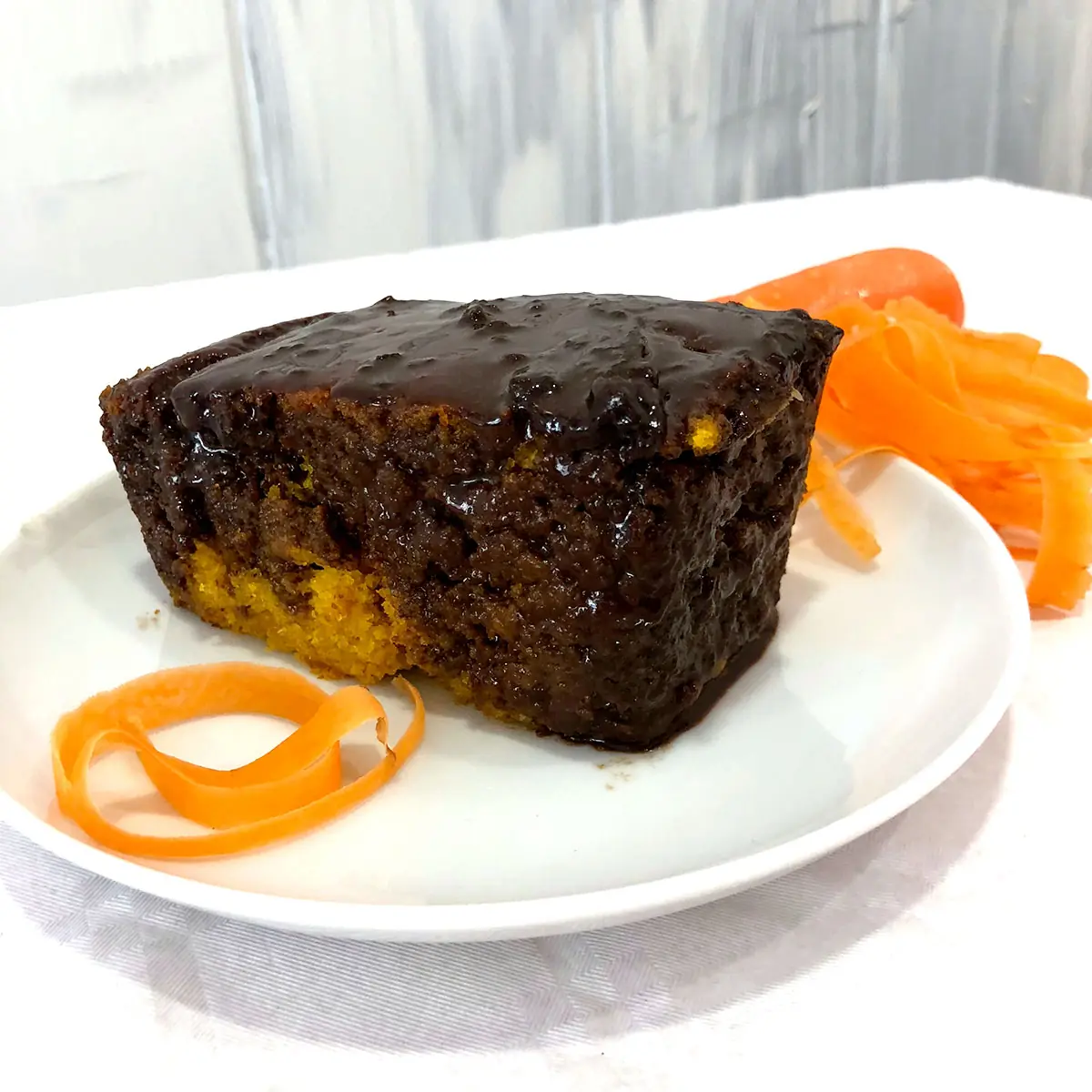 Slice of chocolate-glazed carrot cake with a coconut filling layer, garnished with carrot ribbons on a white plate, at K'Delicia Restaurant Brasileño