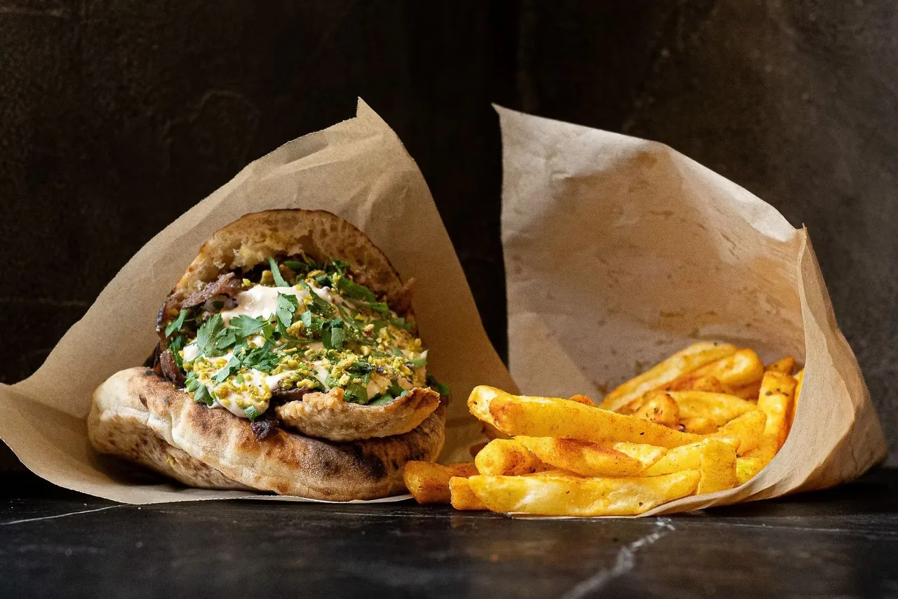 A toasted pita sandwich topped with herbs and crushed nuts wrapped in paper beside a paper cone of fries