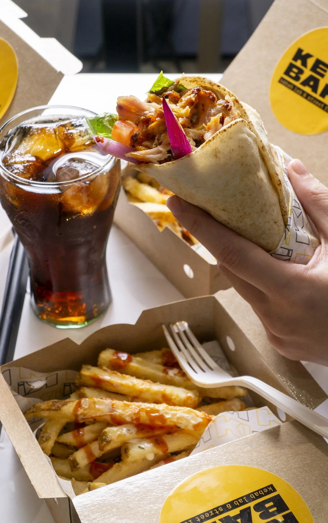 A hand holding a wrapped kebab above a box of fries and a glass of cola