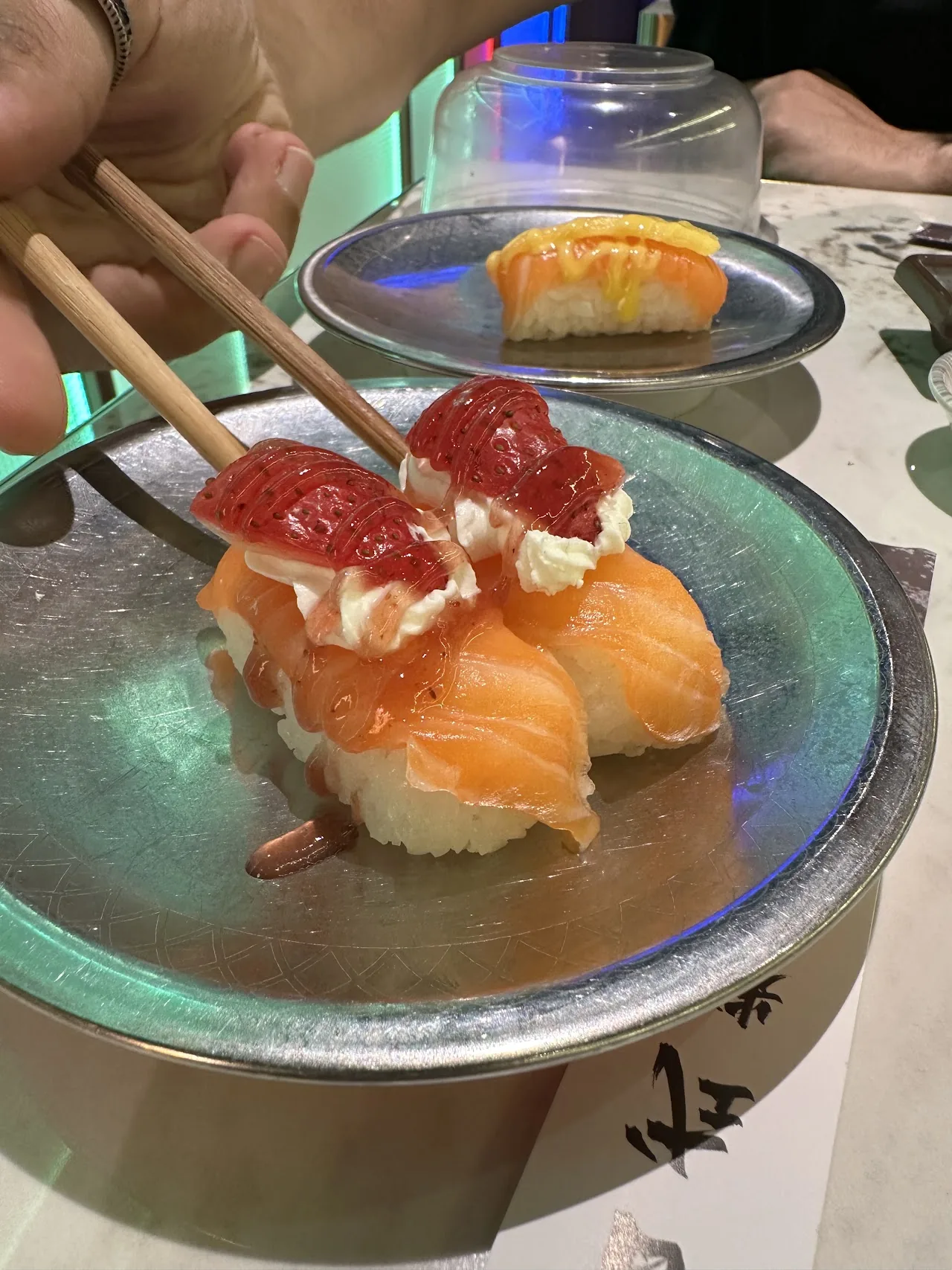 Chopsticks holding a piece of nigiri topped with diced tuna and sauce, more sushi behind