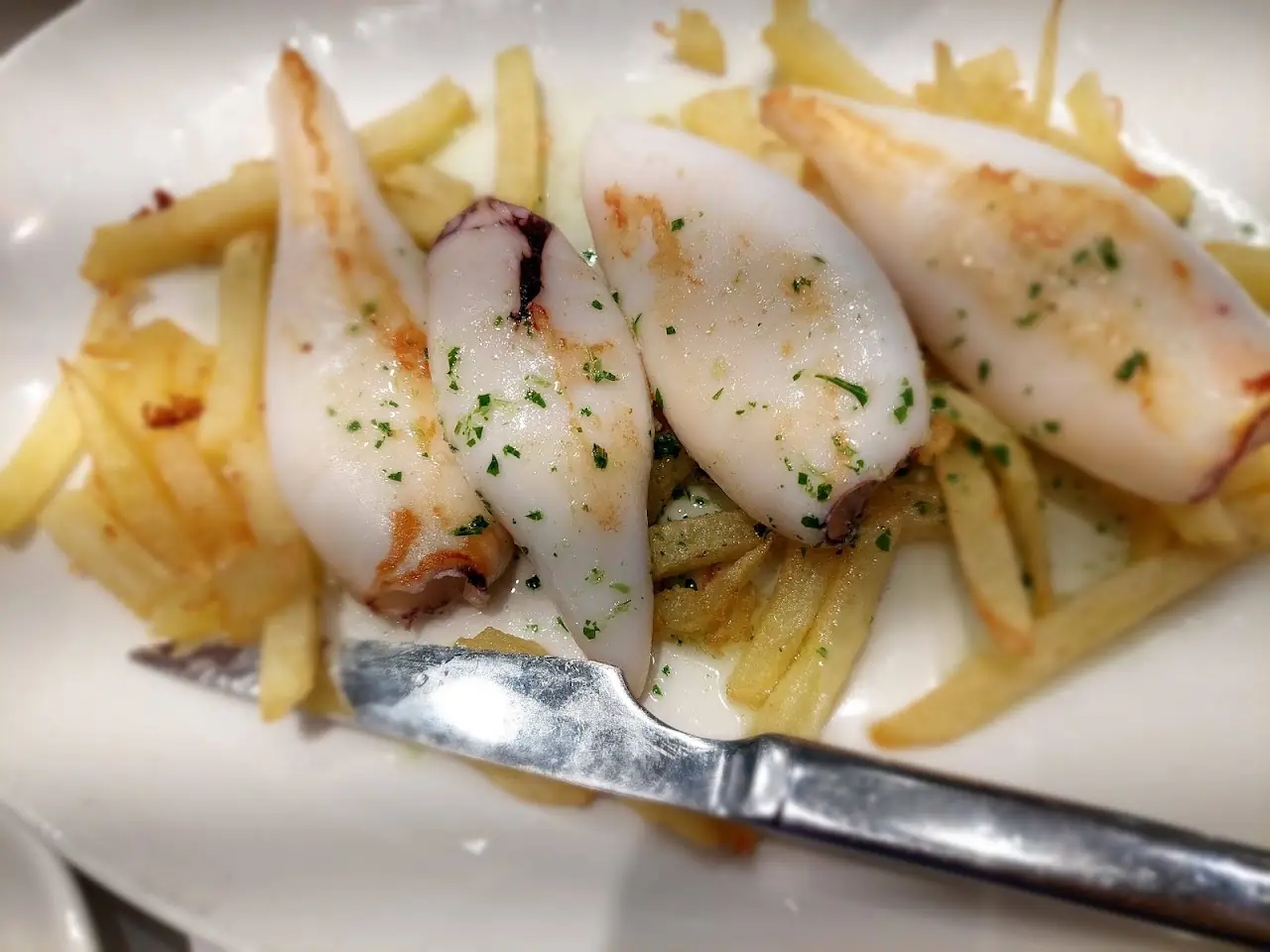 Grilled squid pieces over a bed of fries, with a knife resting on the plate.