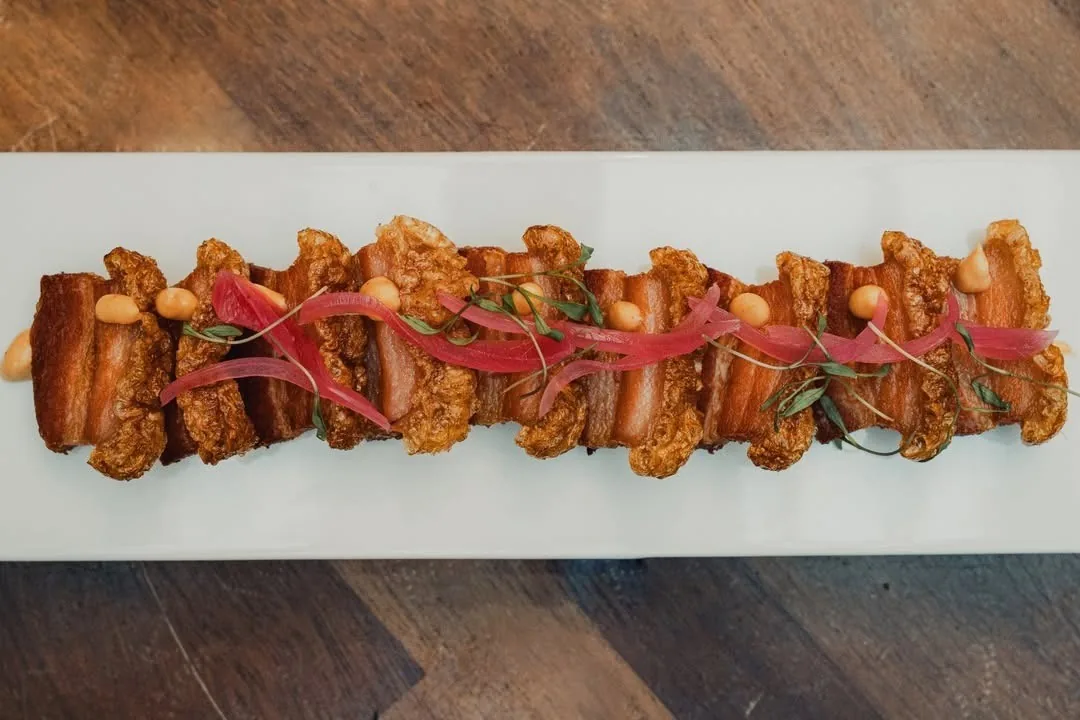 A long white board of fried pork pieces topped with red onion and herbs on a wooden table