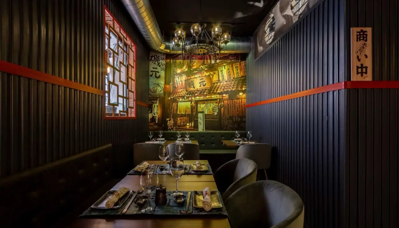 Dark Japanese-themed dining room with a lit izakaya-street mural, globe chandelier, orange trim and set tables with grey chairs