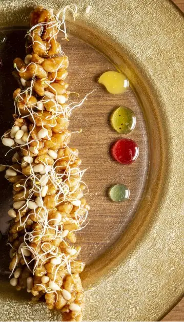Glazed fried batons topped with sprouts and pine nuts on a wooden plate beside three dots of coloured sauce