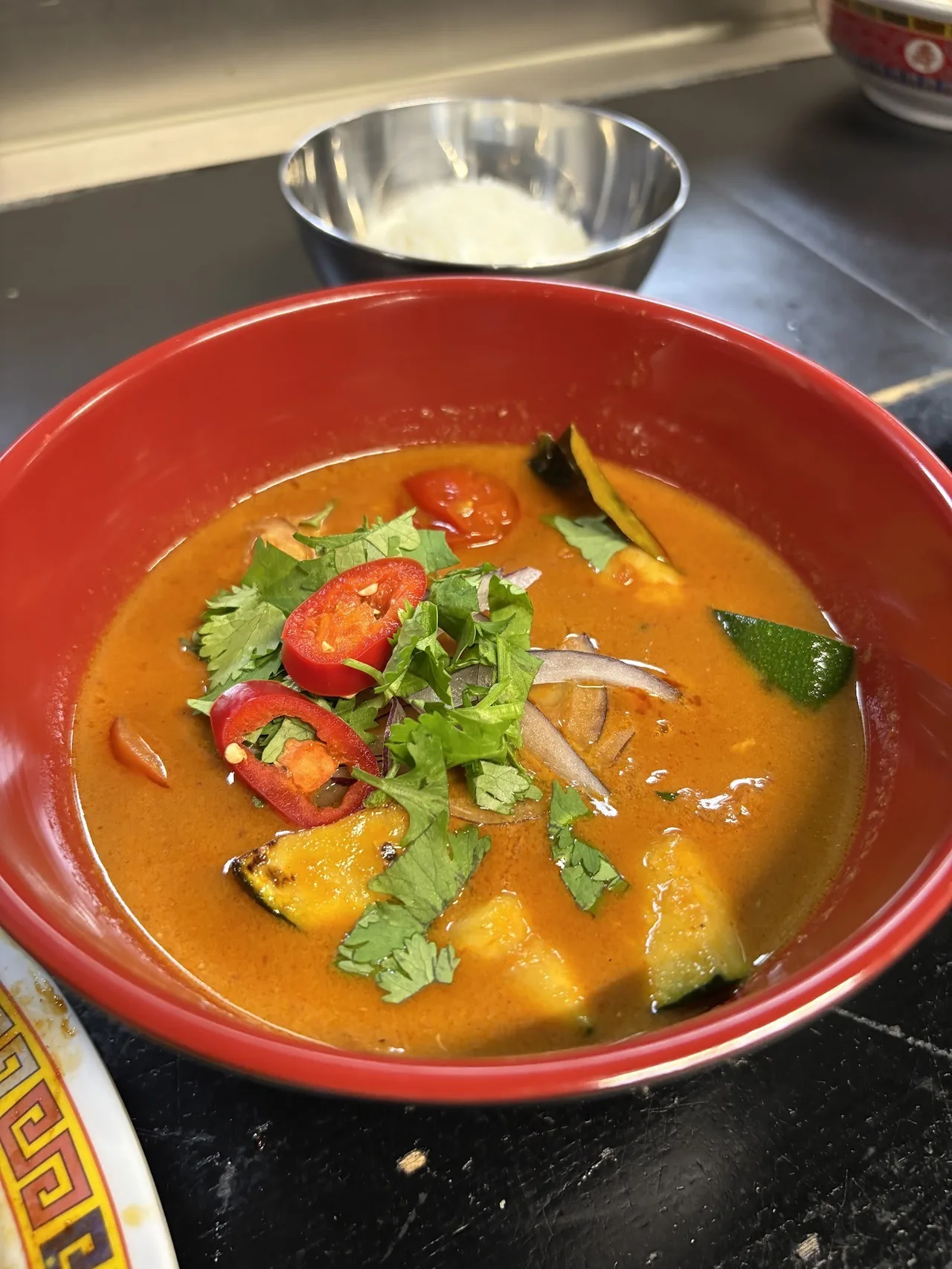 A red bowl of orange curry with pumpkin, red chili and fresh herbs