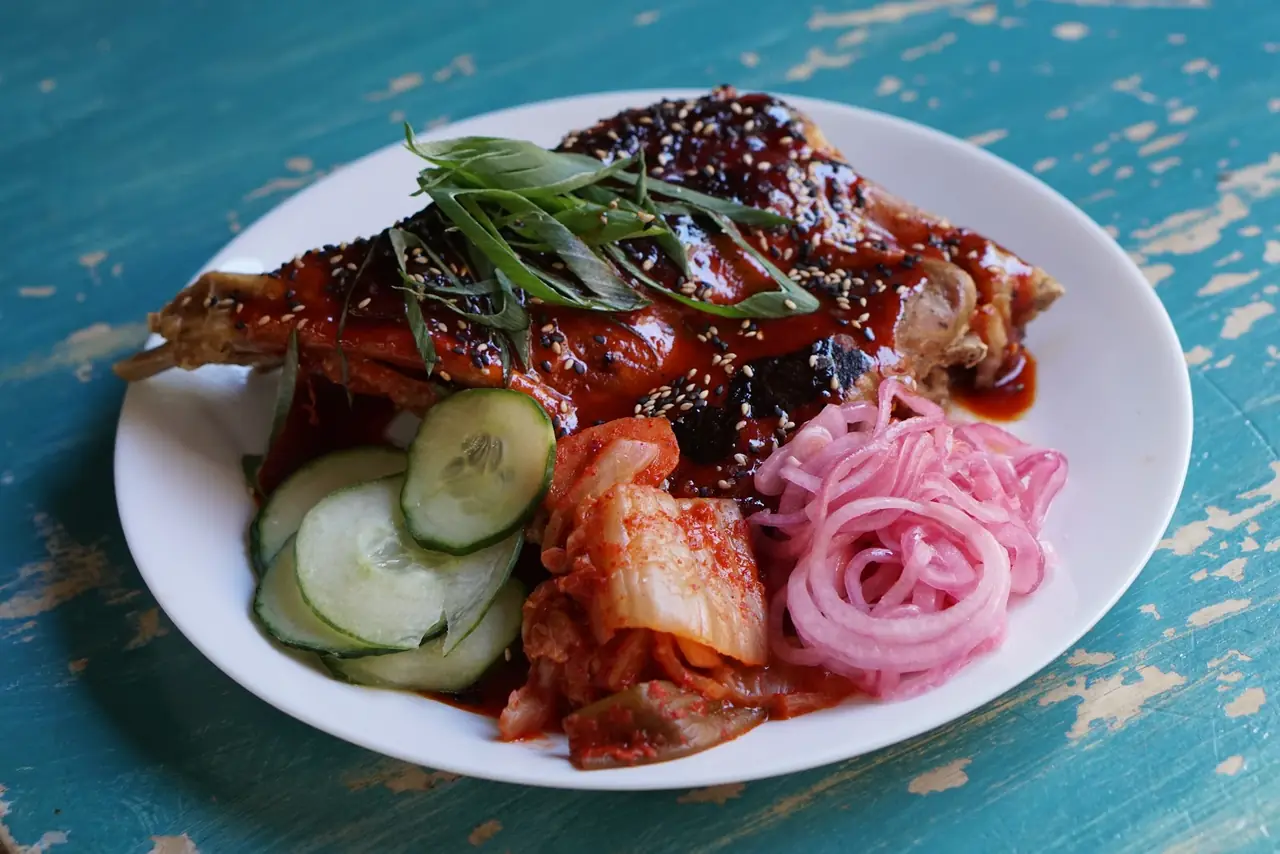 Glazed spicy chicken leg with sesame seeds, served with kimchi, cucumber and pickled red onion