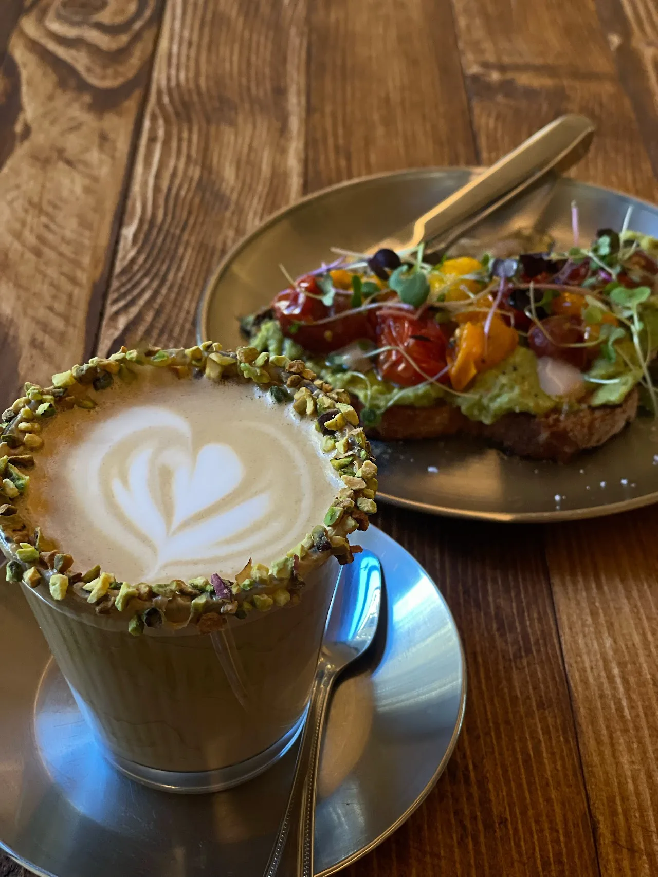 A latte with a pistachio-crusted rim next to a slice of toast topped with tomatoes and greens