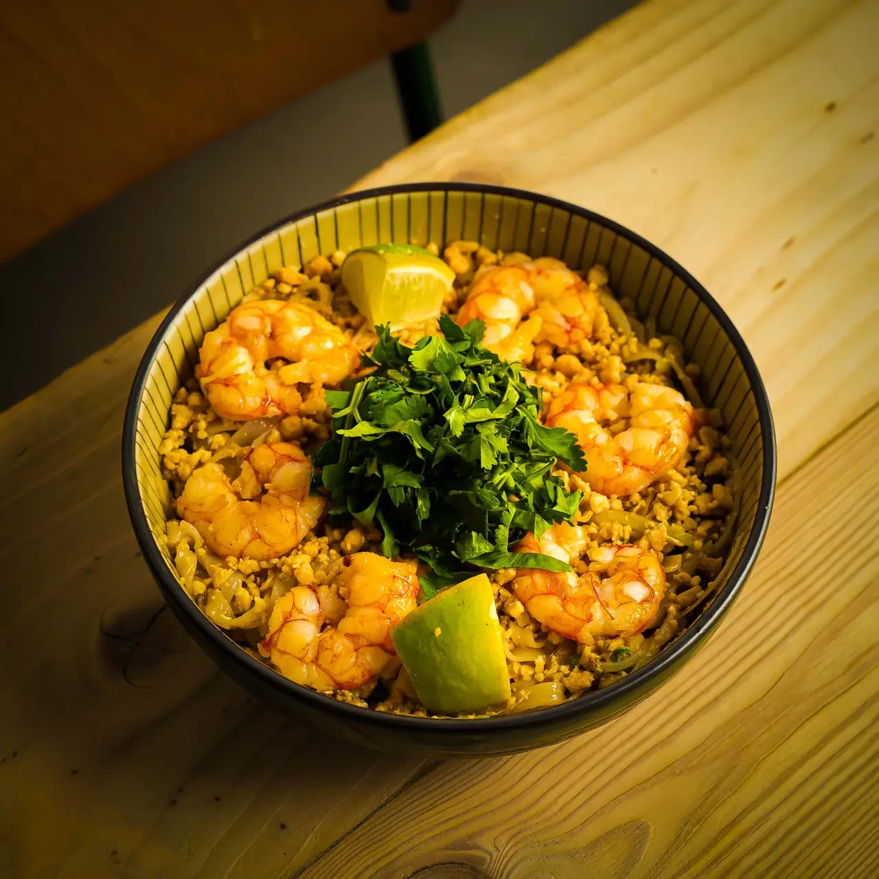 Noodle bowl topped with grilled shrimp, chopped cilantro, and lime wedges on a wooden table at Koku Kitchen