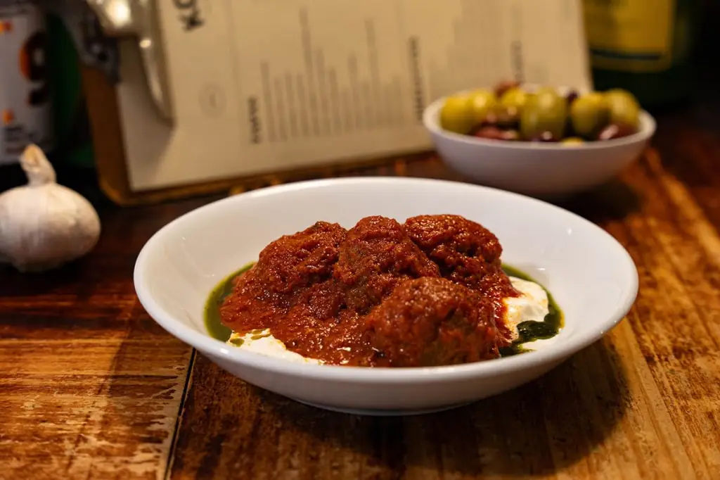 Meatballs in a red sauce over yogurt with green herb oil, in a white bowl at Koska Taverna