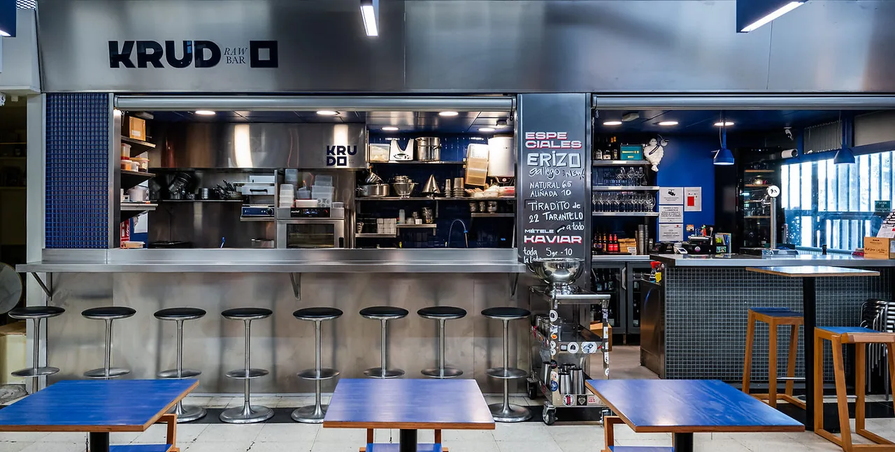 The KRUDO Raw Bar stall front: a long stainless-steel counter with black stools, an open kitchen behind, a handwritten specials board and blue-topped tables in front