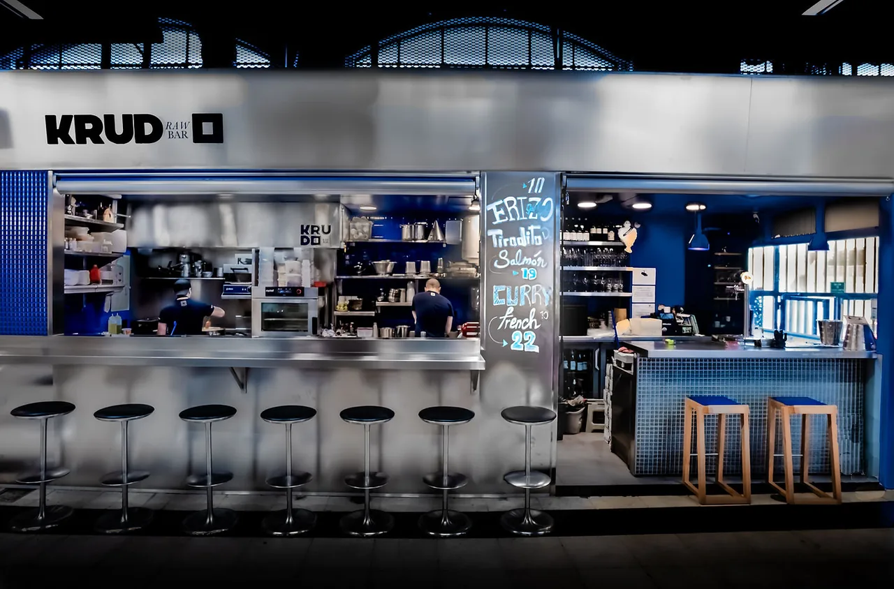 KRUDO Raw Bar at night from across the market hall, steel counter and stools lit blue, two cooks working behind the pass and a chalked specials board between the openings