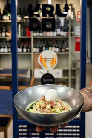 A hand holding out a steel bowl of diced raw fish in pale broth with herb oil and a white quenelle on top, the KRUDO sign and wine shelves behind