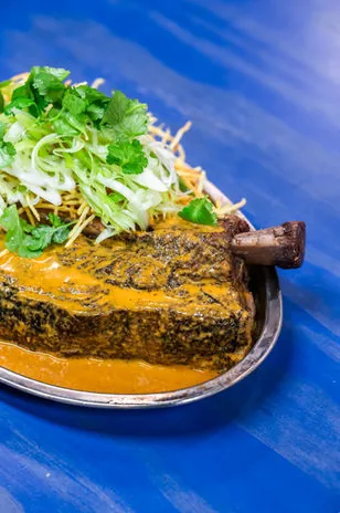 A rib joint under thick orange sauce on a metal plate with shoestring fries, shaved spring onion and coriander, on a bright blue table