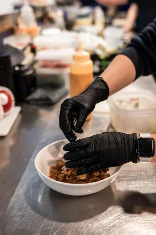 Black-gloved hands plating shredded braised meat into a white bowl on a steel kitchen pass, squeeze bottles behind