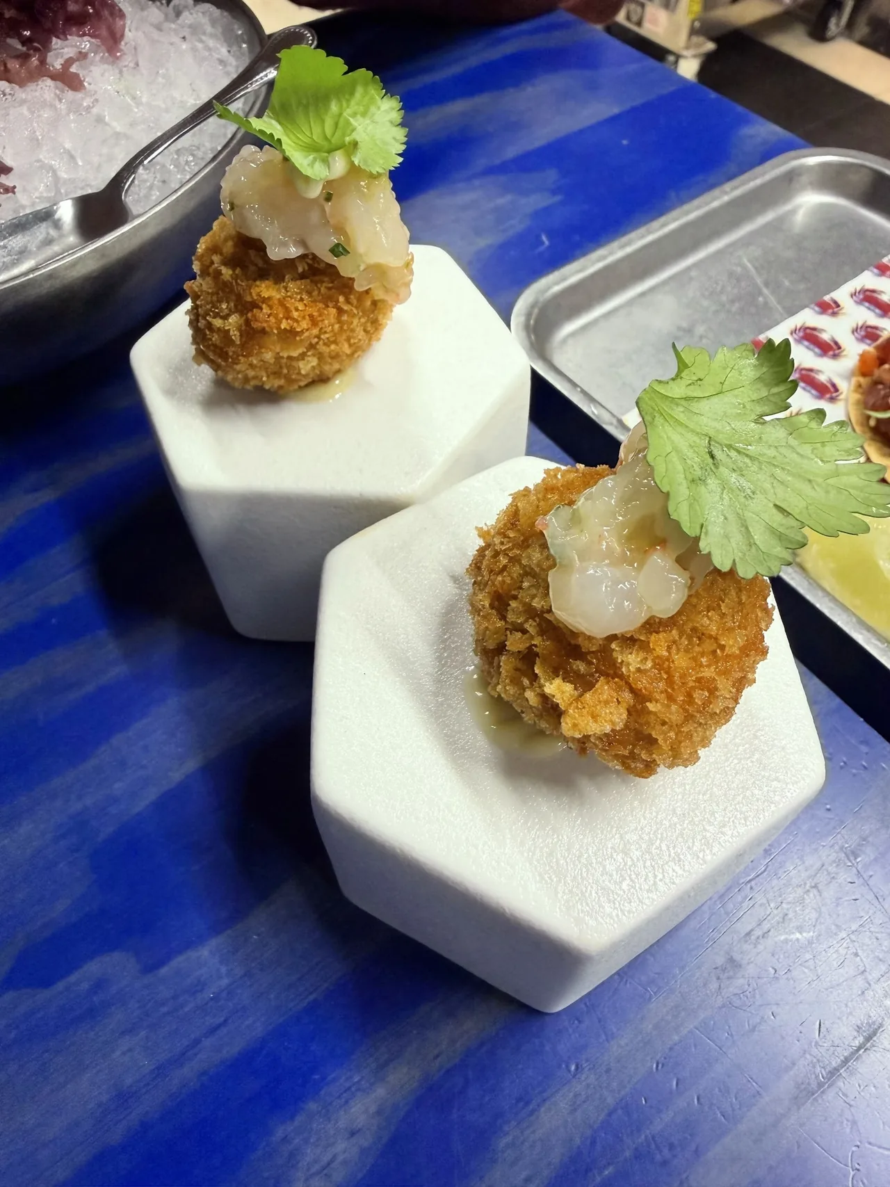 Two breadcrumbed croquetas topped with chopped raw prawn and a coriander leaf on white hexagonal stands