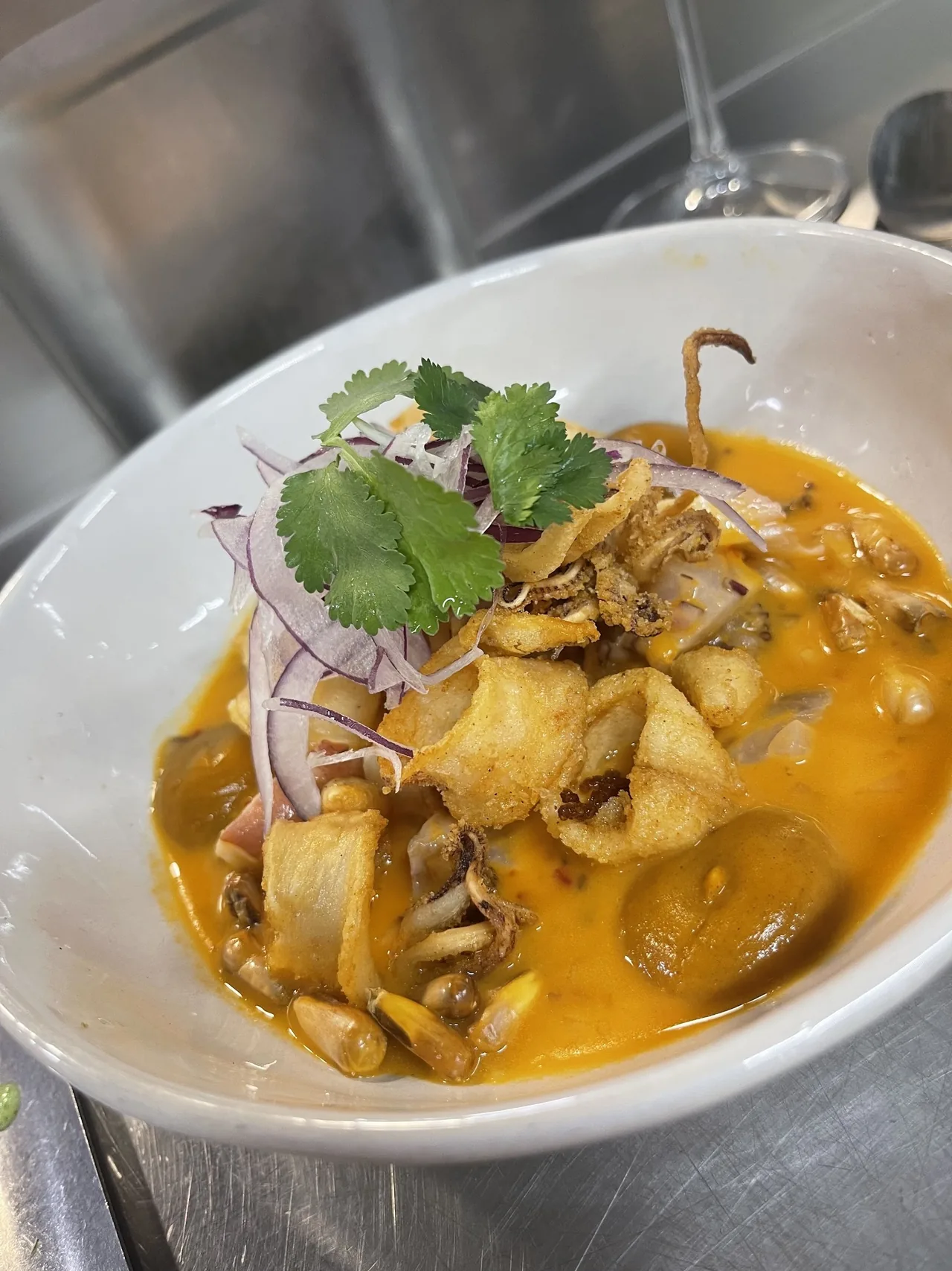 A white bowl of seafood in orange broth topped with fried squid, red onion and coriander