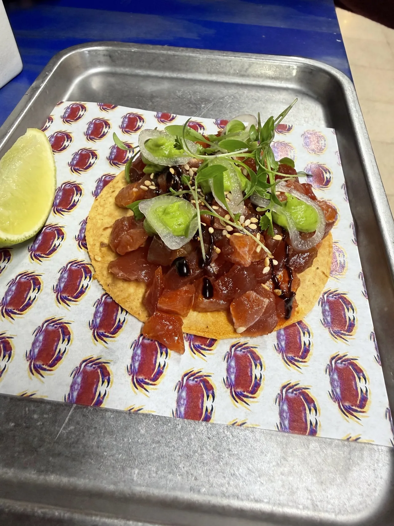 A tostada topped with diced raw tuna, green sauce and sprouts on a metal tray