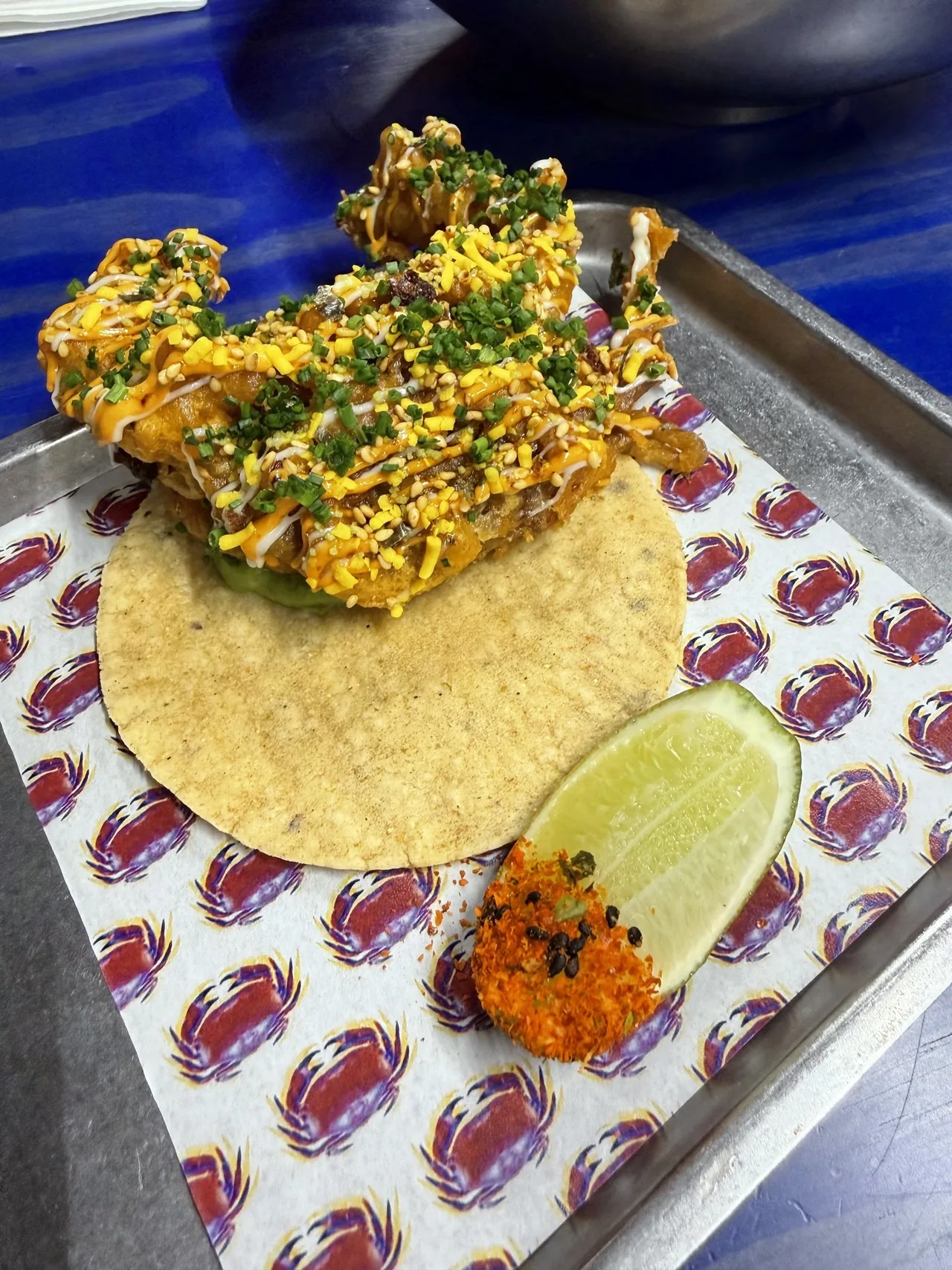 A tortilla topped with a fried soft-shell crab, sauce and chives beside a lime wedge