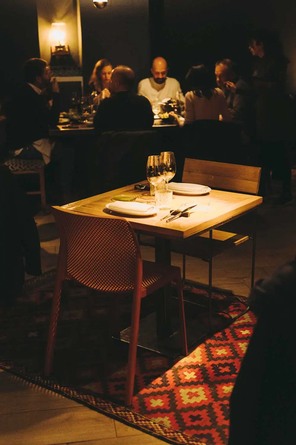 A low-lit dining room: a small pale wood table laid for two with wine glasses, white napkins and a ribbed tumbler, a rust-red moulded chair and a wooden bench beside it, standing on a red and cream kilim runner over slate tiles, with a full table of diners behind.