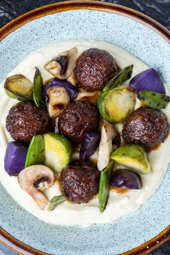 A pale blue bowl of dark meatballs with roasted vegetables on a white puree